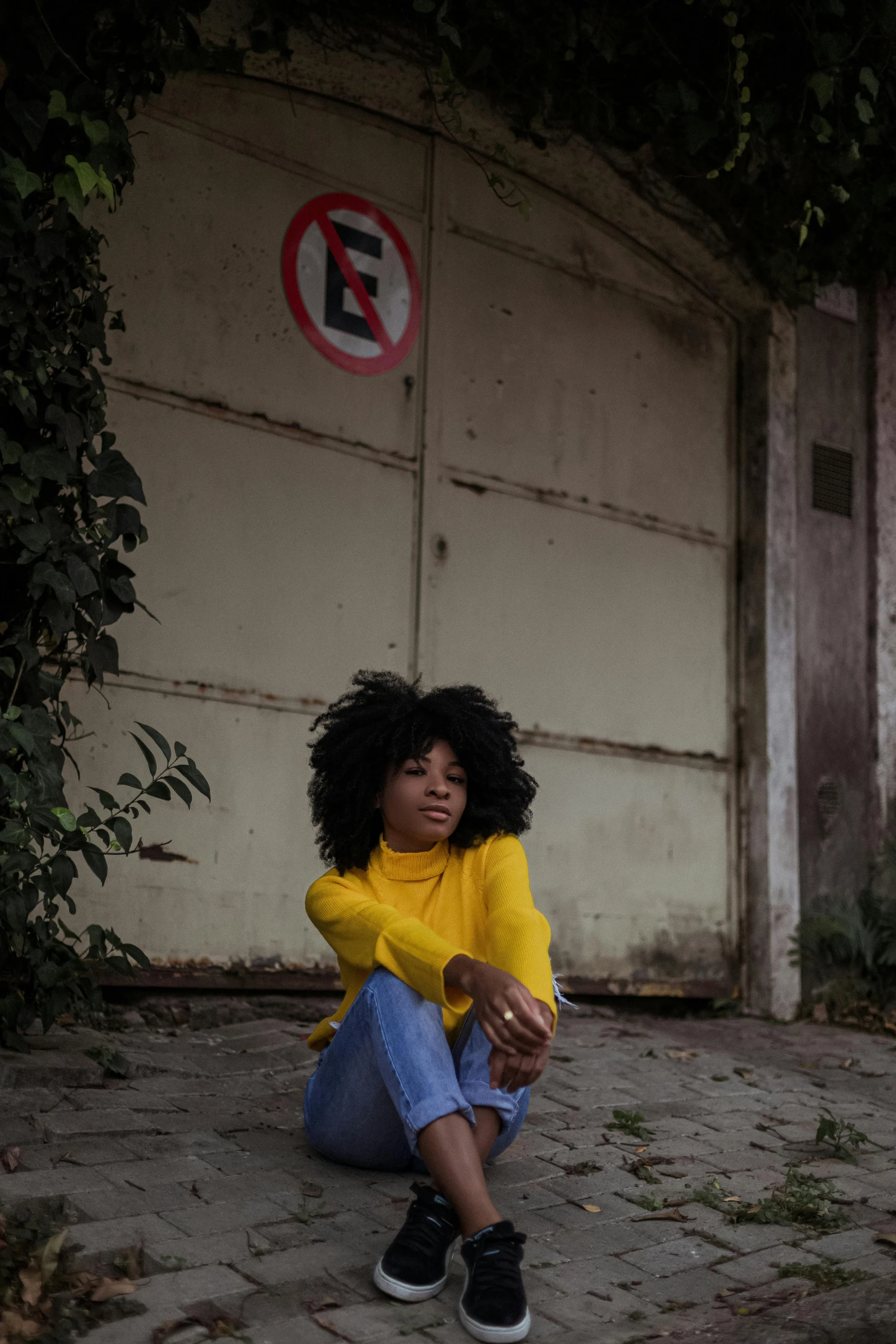 a woman sitting on the ground in front of a garage, by Lily Delissa Joseph, pexels contest winner, wavy hair yellow theme, afro, sza, in sao paulo