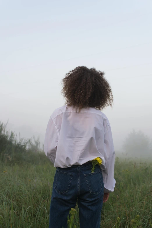 a woman standing on top of a lush green field, an album cover, trending on pexels, detailed hair foggy, white shirt and jeans, human staring blankly ahead, back light