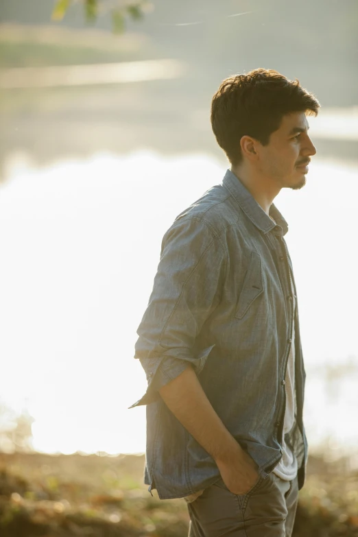 a man standing in front of a body of water, an album cover, inspired by John Luke, looking away from camera, backlighting, mark edward fischbach, wearing a linen shirt