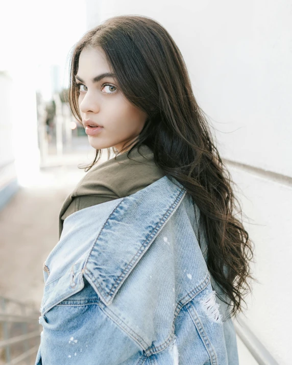 a woman in a denim jacket leaning against a wall, trending on pexels, portrait sophie mudd, wearing shoulder cape, color image, fair olive skin
