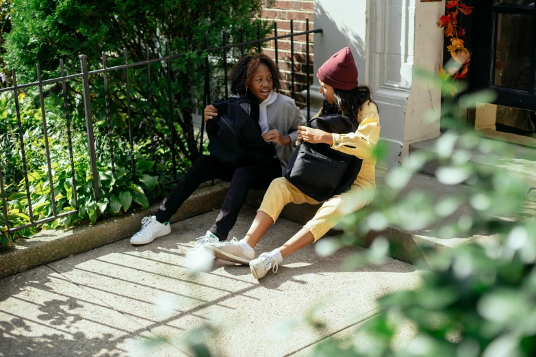 a couple of people sitting next to each other on a sidewalk, by Nina Hamnett, pexels contest winner, happening, black and yellow tracksuit, woman holding another woman, at home, high school girls