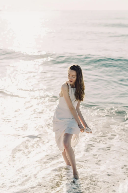 a woman standing on top of a beach next to the ocean, wearing a white dress, pale skin, splashing, softly - lit