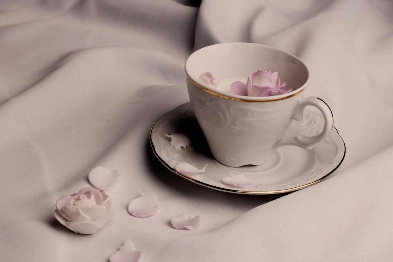 a white cup sitting on top of a saucer, a still life, inspired by Annie Rose Laing, unsplash, romanticism, rose petals, soft lilac skies, white splendid fabric, miniature porcelain model