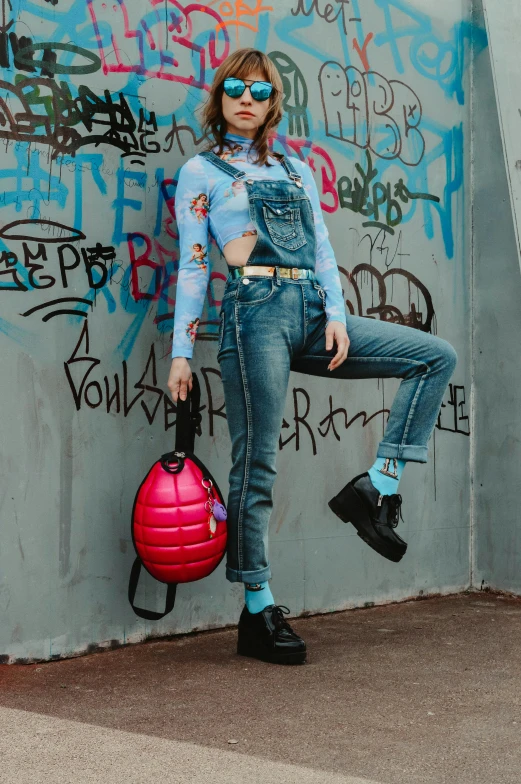 a woman leaning against a wall with graffiti on it, trending on pexels, blue overalls, wearing strawberry backpack, full body with costume, ad image