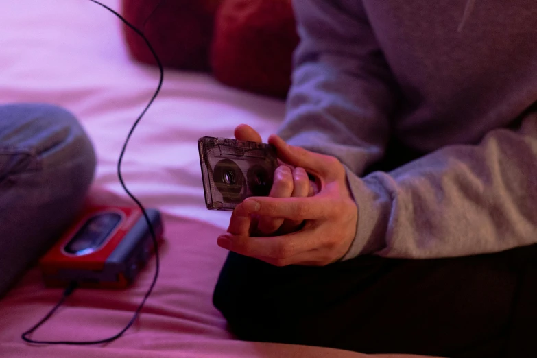 a person sitting on a bed holding a camera, an album cover, trending on pexels, vcr tape, purple ambient light, playing games, cassette tape