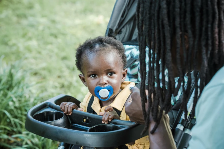 a little girl sitting in a stroller with a pacifier in her mouth, pexels contest winner, man is with black skin, walking at the garden, community celebration, avatar image