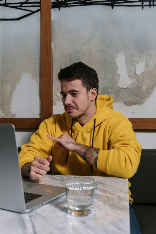 a man sitting at a table in front of a laptop computer, a colorized photo, trending on pexels, realism, yellow clothes, scolding, liam, trending on r/streetwear