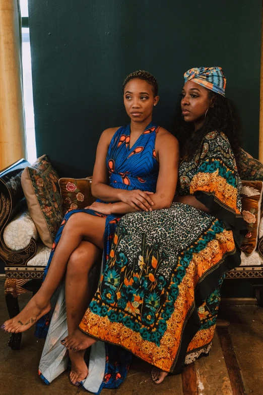 two women sitting on a couch in a room, a portrait, by Ella Guru, unsplash, renaissance, wearing an african dress, modeling for dulce and gabanna, “ full body, promotional image