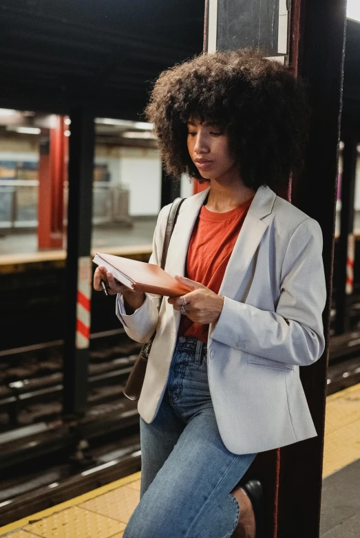 a woman sitting on a train platform reading a book, a portrait, by Carey Morris, pexels contest winner, wearing a blazer, with afro, orange line, standing