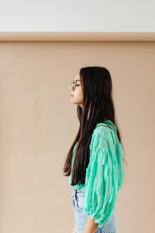 a woman standing in front of a beige wall, inspired by Elsa Bleda, trending on pexels, green robe, asian girl with long hair, teenager girl, girl with glasses