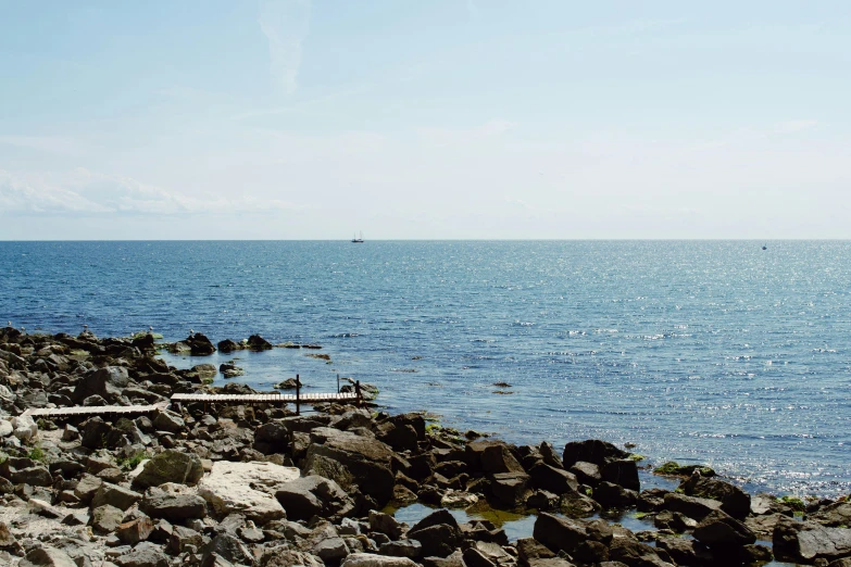 a bench sitting on top of a rocky beach next to the ocean, a picture, unsplash, mingei, helsinki, clear blue skies, sailboats in the water, 2000s photo