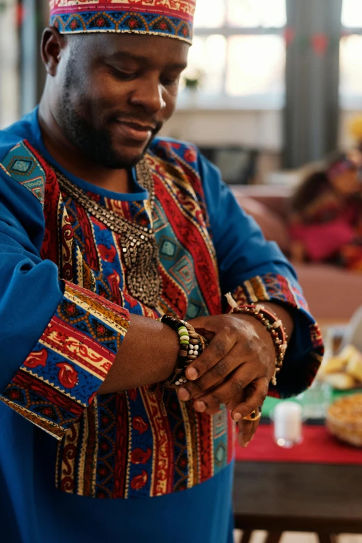 a man standing in front of a table with food on it, inspired by Ras Akyem, patterned clothing, beads, closeup of arms, bringing people together