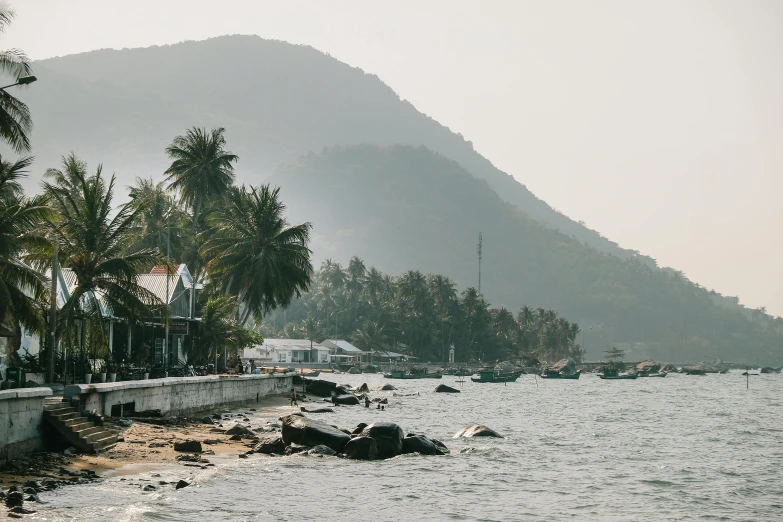 a group of people standing on a beach next to a body of water, vietnam, waterfront houses, near a jetty, mountains and oceans