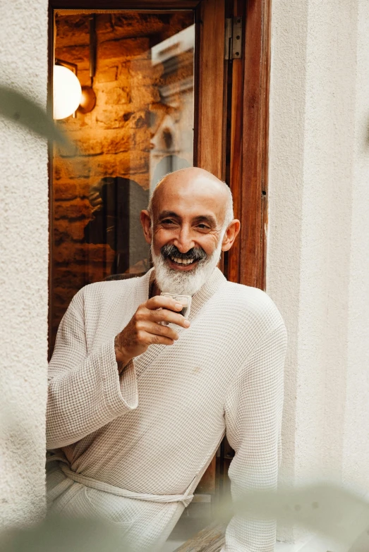 a man with a beard standing in a doorway, inspired by Agnolo Gaddi, pexels contest winner, bald head and white beard, attractive man drinking coffee, happily smiling at the camera, wearing white clothes