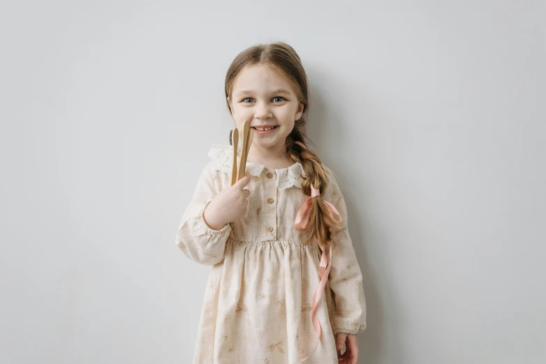 a little girl standing in front of a white wall, by Emma Andijewska, pexels contest winner, cutlery, beige and gold tones, smiling sweetly, peasant dress