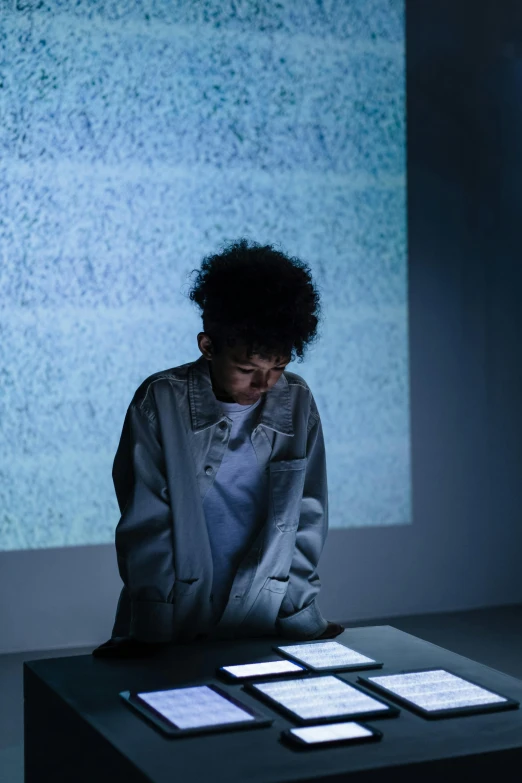 a woman sitting at a table in front of a projector screen, inspired by Ryoji Ikeda, trending on unsplash, interactive art, black teenage boy, sad expression, full body backlight, young boy