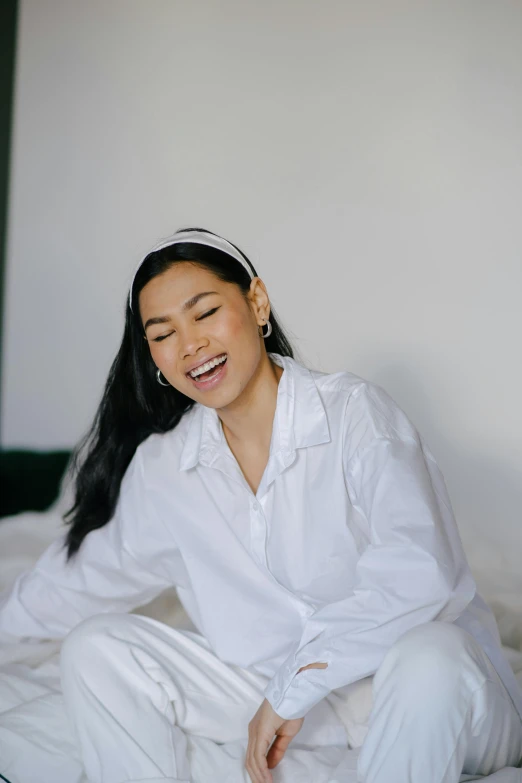 a woman sitting on a bed with her eyes closed, inspired by Ruth Jên, trending on pexels, happening, earing a shirt laughing, glossy white, asian human, low quality photo