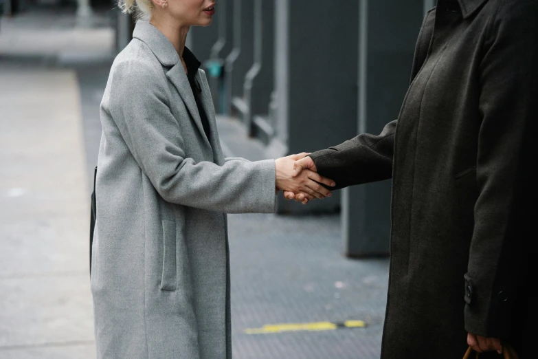 a man and a woman shaking hands on a sidewalk, by Emma Andijewska, pexels contest winner, wearing a duster coat, two different characters, wearing black grey suit, blonde women