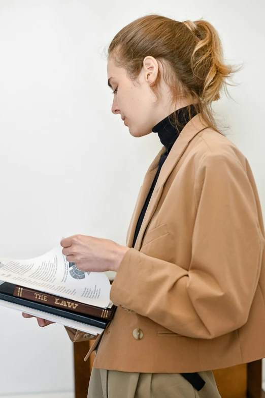 a woman standing in front of a white wall holding a book, light brown coat, law aligned, inspect in inventory image, wearing white sneakers