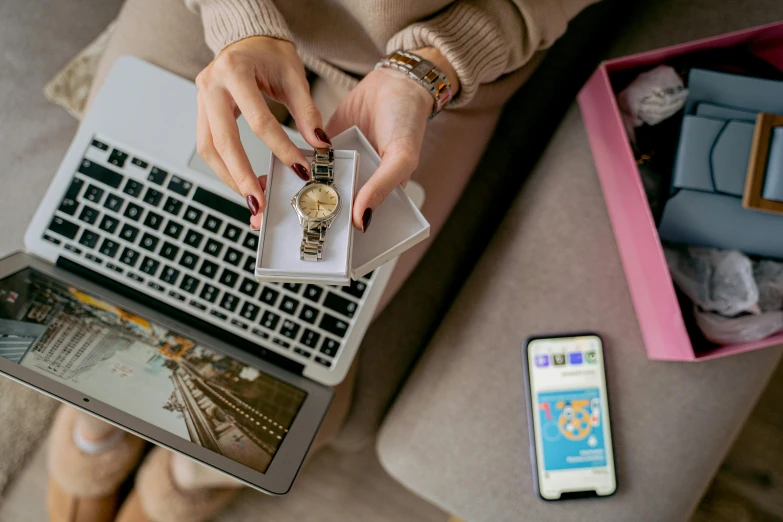 a woman sitting on a couch with a laptop and cell phone, by Julia Pishtar, trending on pexels, holding gold watch, giving gifts to people, sitting on a desk, thumbnail