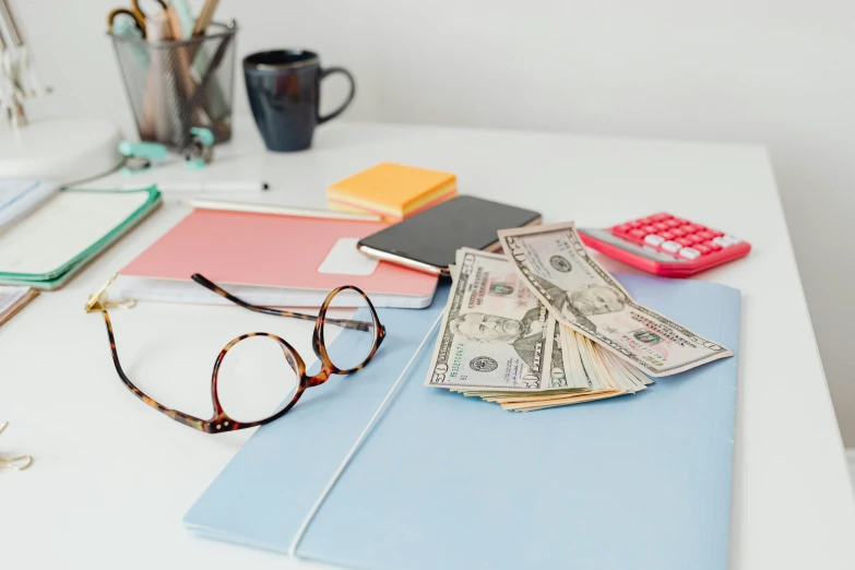 a pile of money sitting on top of a desk next to a calculator, trending on pexels, blue rimmed glasses, pink and red color scheme, thumbnail, table in front with a cup