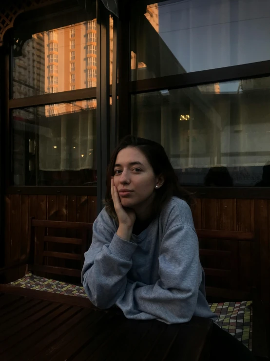 a woman sitting at a table in front of a window, a picture, inspired by Elsa Bleda, pexels contest winner, realism, wearing a grey hooded sweatshirt, in moscow centre, 🤤 girl portrait, at the terrace