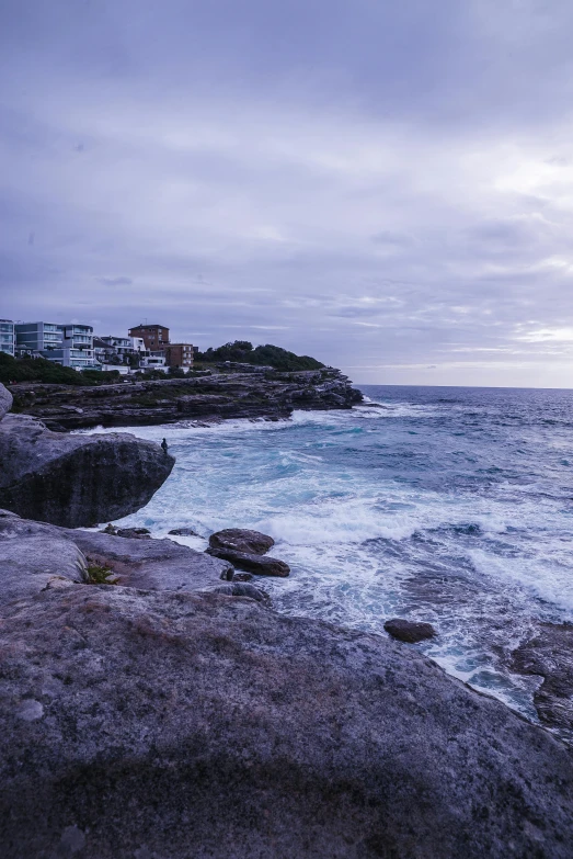 a man sitting on top of a rock next to the ocean, inspired by Sydney Carline, unsplash, australian tonalism, purple, bondi beach in the background, evening storm, slide show