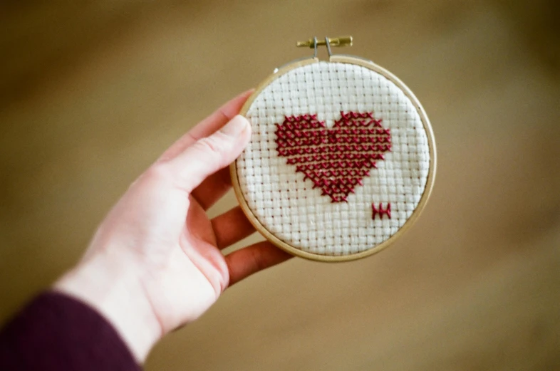 a close up of a person holding a cross stitch heart, by Sylvia Wishart, pexels, maroon, a round minimalist behind, display”, assembled