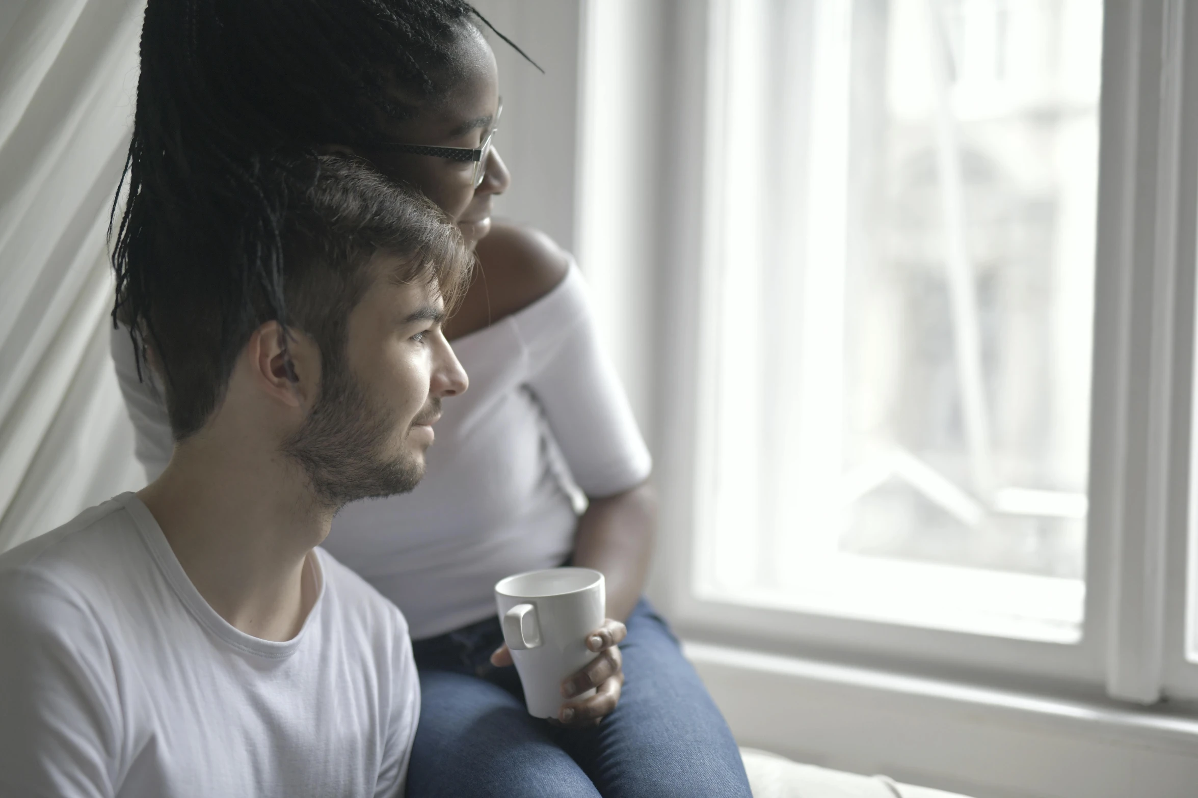 a man and a woman sitting next to each other on a bed, trending on pexels, sitting on a window sill, next to a cup, mixed race, looking across the shoulder