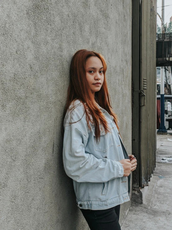 a woman leaning against the side of a building, by Robbie Trevino, jean jacket, a redheaded young woman, low quality photo, trending on r/streetwear