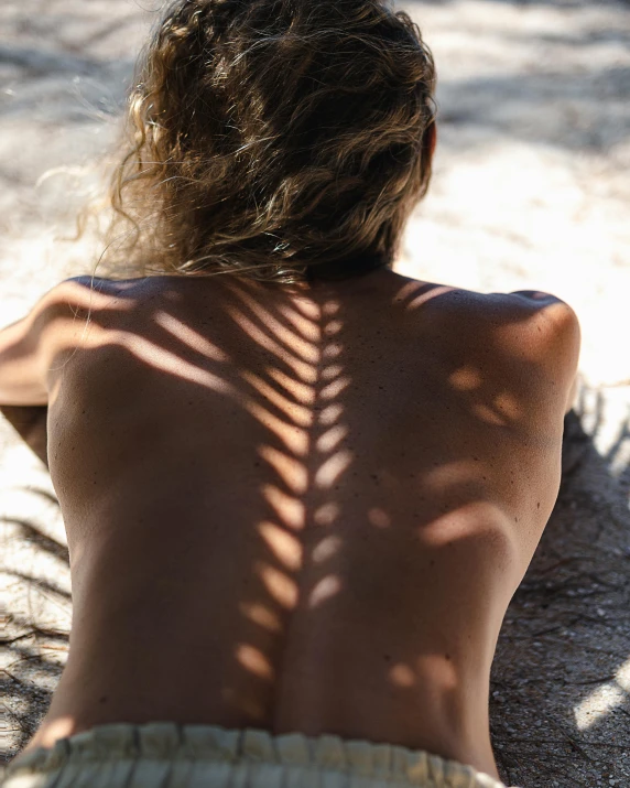 a woman sitting on top of a sandy beach, a tattoo, inspired by Max Dupain, trending on unsplash, naturalism, trees cast shadows on the wall, human spine, in a sunbeam, arched back