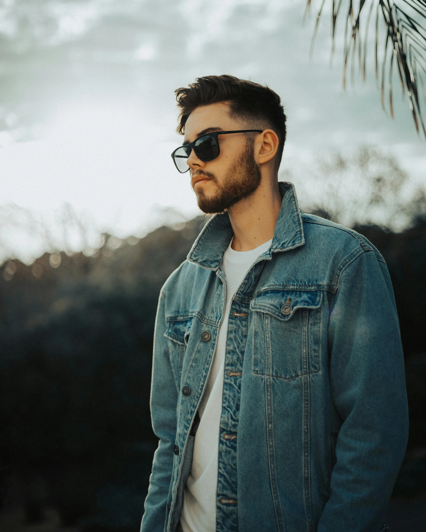 a man standing in front of a palm tree, trending on pexels, denim jacket, non binary model, glasses, profile image