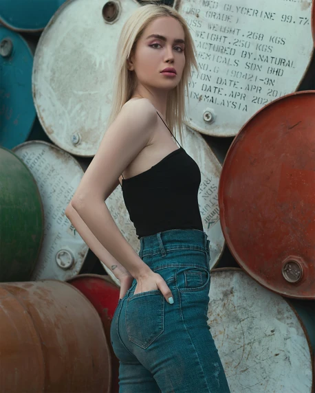 a woman standing in front of a wall of barrels, a colorized photo, inspired by Elsa Bleda, pexels contest winner, portrait of kim petras, ( ( ( wearing jeans ) ) ), anna nikonova aka newmilky, detailed product image