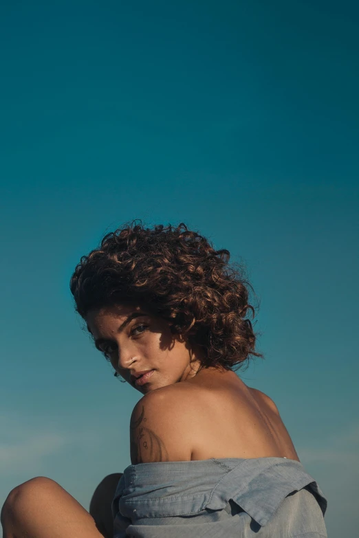 a woman sitting on top of a sandy beach, an album cover, by Alexis Grimou, trending on pexels, renaissance, curly pixie hair, clear blue sky, back light, halfbody headshot