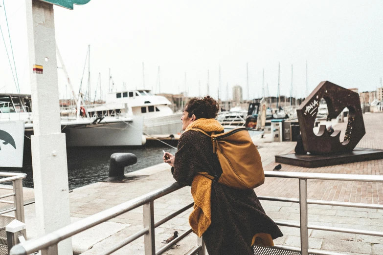 a woman standing on a pier looking at boats, pexels contest winner, happening, wearing a brown cape, wearing a yellow hoodie, the man have a backpack, female streetwear blogger