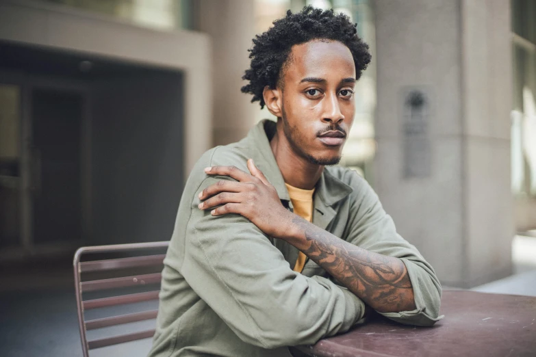 a man sitting at a table with his arms crossed, inspired by Afewerk Tekle, pexels contest winner, playboi carti portrait, calm weather, thin aged 2 5, loosely cropped