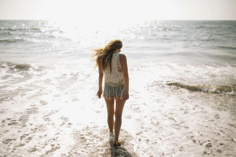 a woman standing on top of a beach next to the ocean, pexels, walking away, wearing a camisole and shorts, single vague light, wet skin and windblown hair