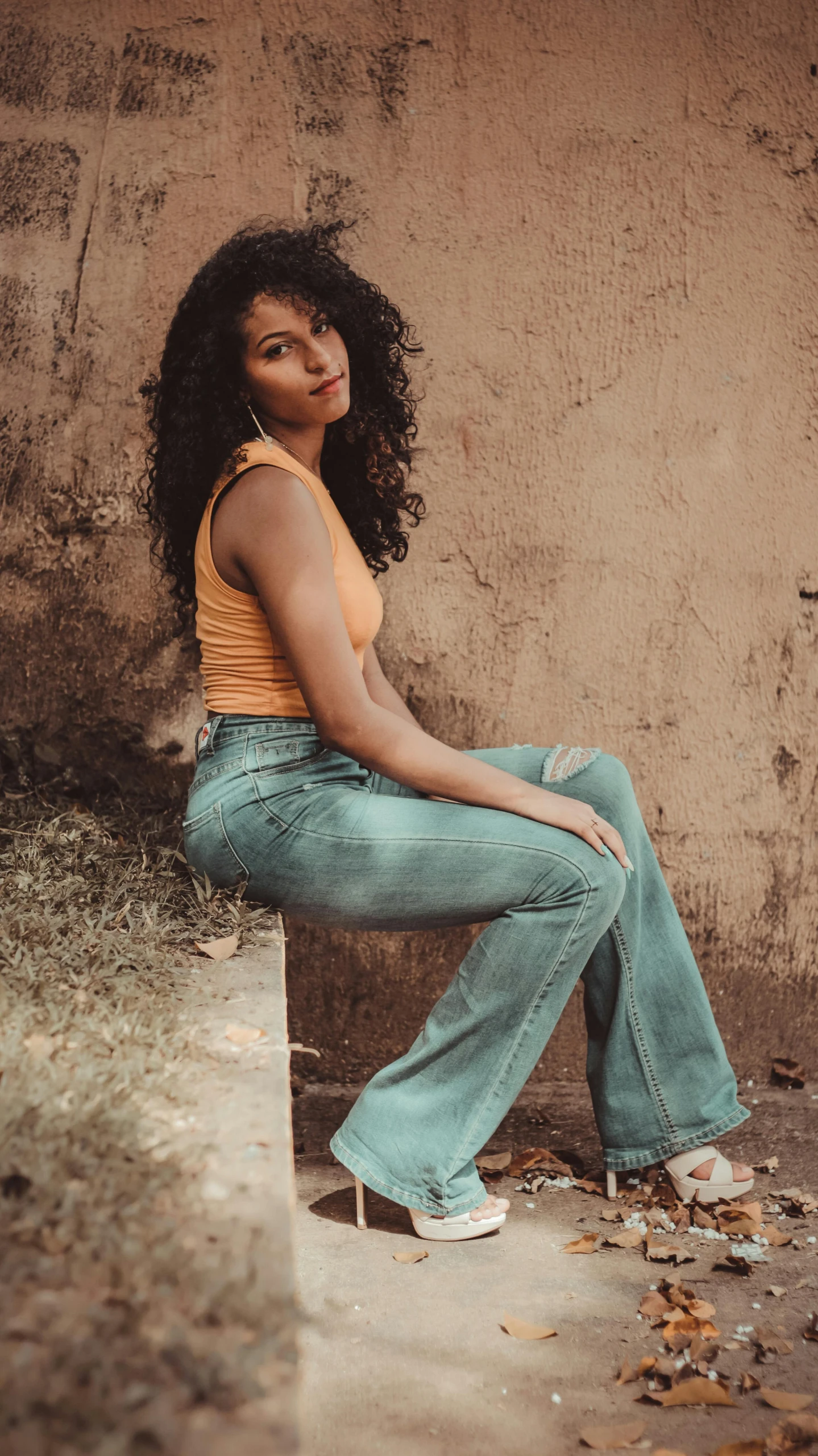 a woman sitting on a ledge in front of a wall, an album cover, inspired by Elsa Bleda, trending on pexels, renaissance, jeans pants, curly afro, wearing yellow croptop, turquoise rust