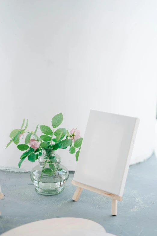 a picture frame sitting on top of a table next to a vase of flowers, a minimalist painting, white wall coloured workshop, white canvas background, indoor setting, romantic greenery