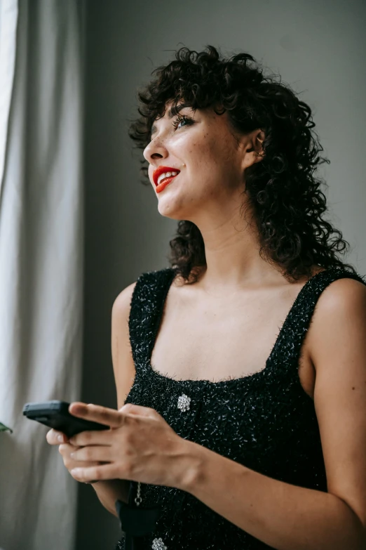 a woman standing in front of a window holding a cell phone, curly dark hair, wearing a black dress, profile image, curly bangs