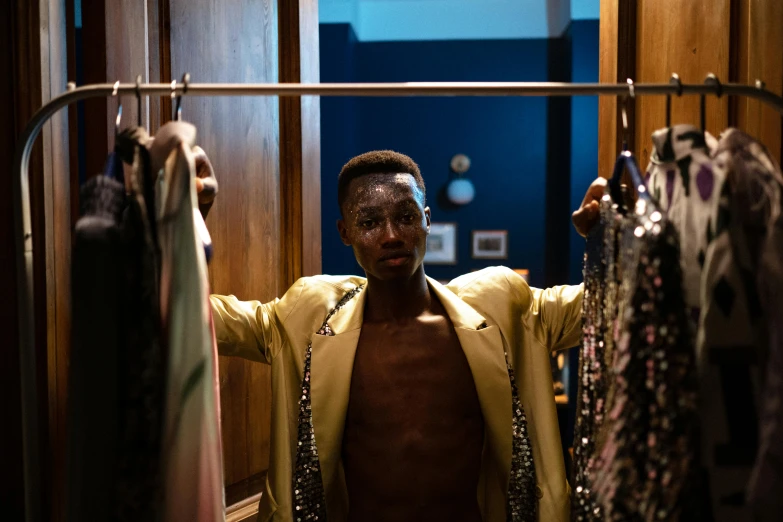 a man standing in front of a rack of clothes, by Julia Pishtar, happening, grace jones, blue gold suit, theater dressing room, sparkly