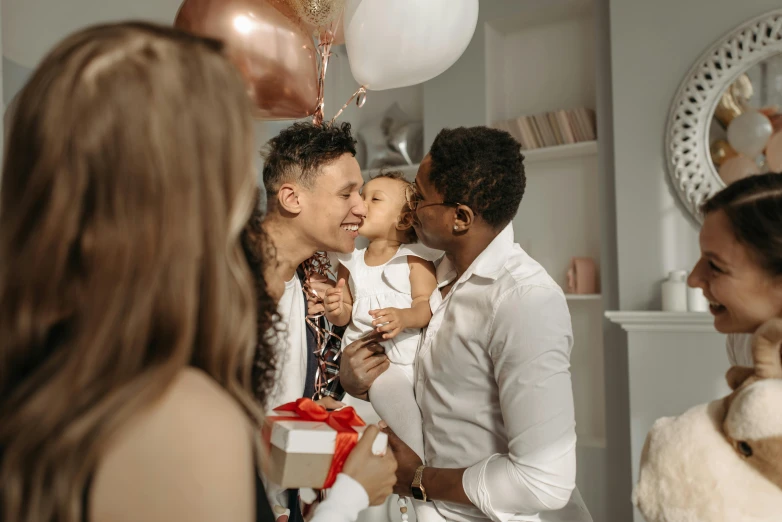 a group of people standing around a birthday cake, father holds child in the hand, white and gold color scheme, couple kissing, man holding a balloon