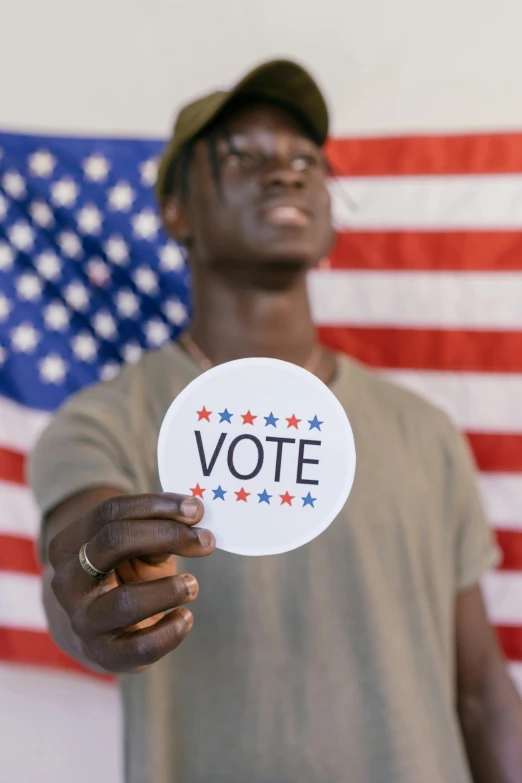 a man holding a vote sticker in front of an american flag, a picture, by Carey Morris, shutterstock, renaissance, man is with black skin, instagram story, model posing, 🚿🗝📝