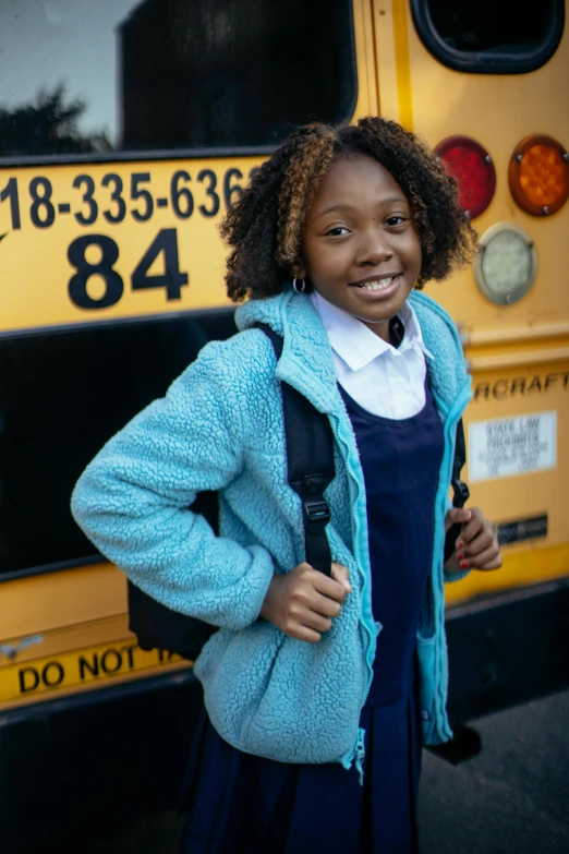 a young girl standing in front of a school bus, wearing a blue jacket, dark skinned, promo image, multiple stories