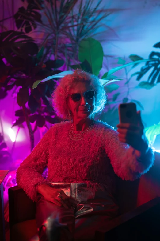 a woman sitting in a chair holding a cell phone, an album cover, by Matt Cavotta, pexels, elderly woman, night clubs and neons, taking a selfie, biophilia