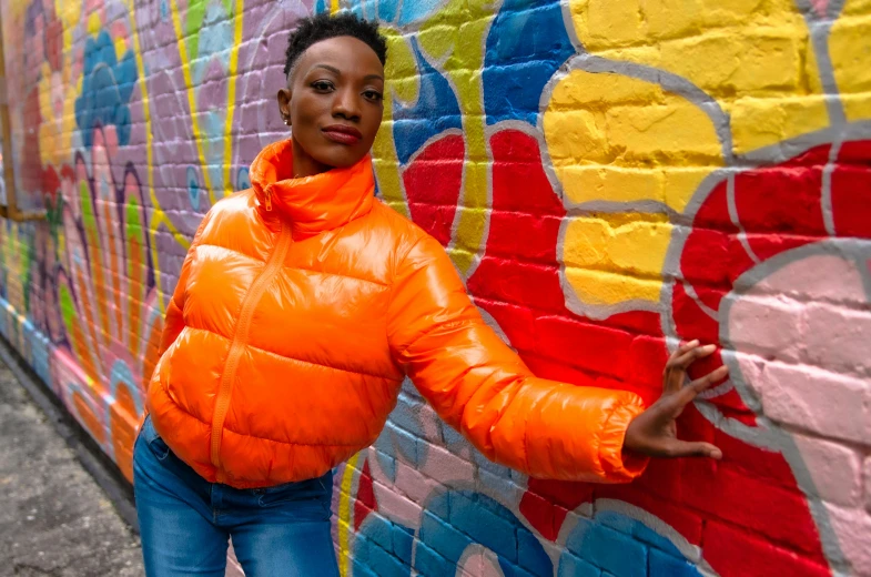 a woman standing in front of a colorful wall, graffiti, orange reflective puffy coat, striking a pose, headshot, maria borges