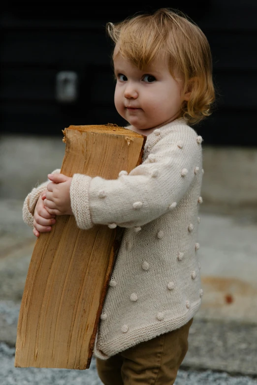 a little girl holding a piece of wood in her hands, an album cover, by David Simpson, unsplash, cardigan, natural materials, holding a giant book, bumpy skin