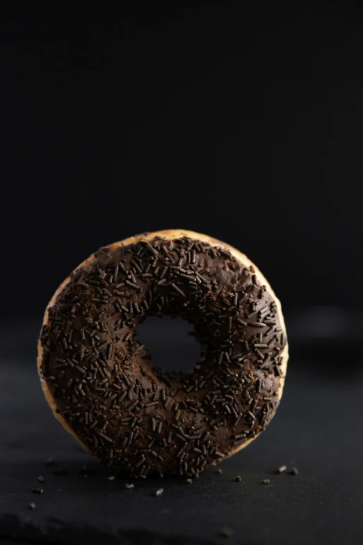 a chocolate donut with sprinkles on a black surface, a portrait, unsplash, detailed product image, 3/4 front view, chile, made of tar