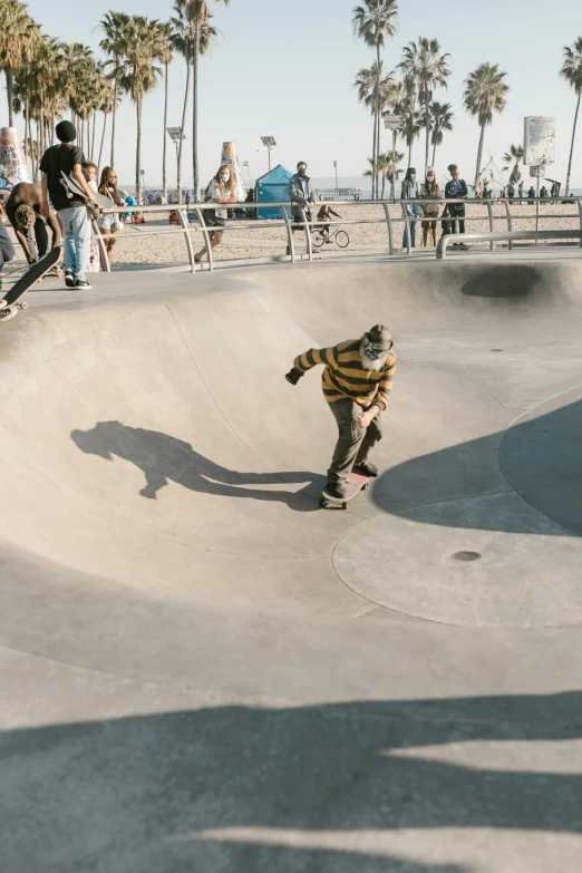 a man riding a skateboard up the side of a ramp, sand swirling, bowl, people walking around, los angelos