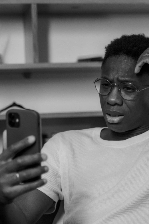 a man sitting on a couch holding a cell phone, a black and white photo, by Dean Ellis, pexels, realism, young thug, wearing black frame glasses, looking in mirror, black teenage boy